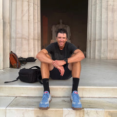 A man wearing the Mackall 45 Americana sits on marble steps in front of a statue inside a large columned structure, smiling at the camera.