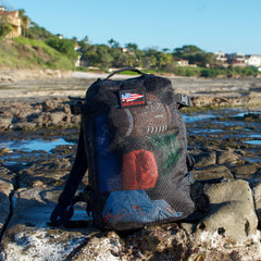 The Mesh Ruck, a black mesh backpack, sits on rocky coastal terrain with houses and greenery behind—ideal as a quick-drying beach bag for all your seaside adventures.