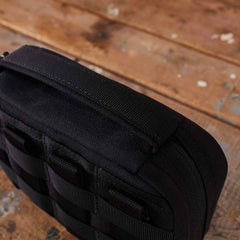 Close-up of the GR1 Field Pocket - USA, a black tactical bag with webbing, perfect as an electronics organizer, resting on a scratched wooden surface.