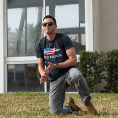 A man wearing sunglasses, a backpack, and the GORUCK Spearhead Tee - Stars & Stripes - Tri-BlendX kneels on grass near a building with glass windows, embodying the spirit of the Special Forces while displaying the iconic GORUCK spearhead on his gear.