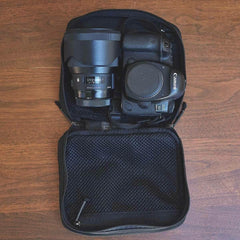 A GR2 Field Pocket by GORUCK, crafted from durable 1000D Cordura, rests on a wooden surface, containing a black Canon camera with an attached lens. The pocket is open, revealing a mesh compartment above the camera, ideal for storing additional accessories and keeping everything neatly organized.