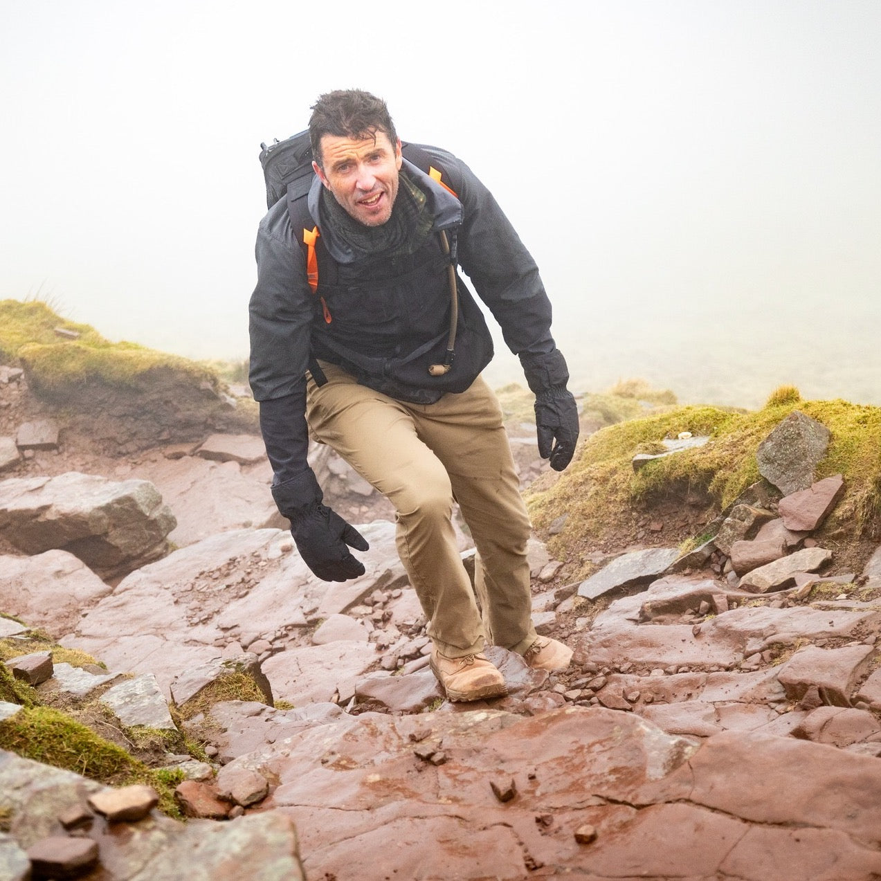 A man in outdoor gear, carrying the Rucker 5.0 rucksack with lumbar support, hikes up a rocky, foggy trail surrounded by moss and grass.
