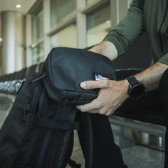 Person wearing a watch attaching a black GORUCK Field Pocket with American flag patch to a black backpack in an indoor seating area