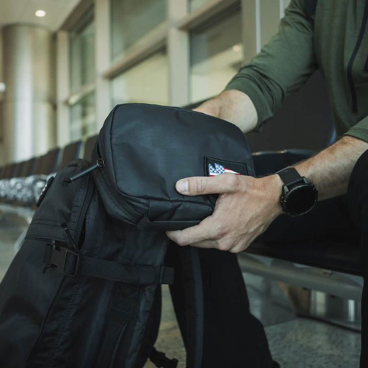 In an airport terminal, a traveler sits holding a sleek black travel bag adorned with a distinctive American flag patch. They are dressed in a green shirt and wear a black smartwatch that perfectly complements their GORUCK GR1 Field Pocket - Dyneema electronics organizer tucked inside.