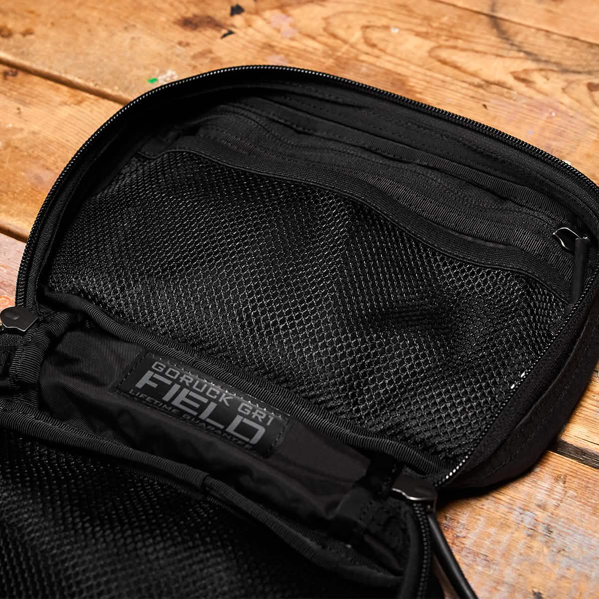 Place the black backpack featuring a mesh pocket on a wooden surface; it serves as an ideal travel accessory. The label displays "GORUCK GR1 Field Pocket - Dyneema," designed with durability for adventure seekers in mind.