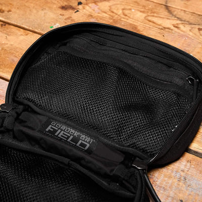 Place the black backpack featuring a mesh pocket on a wooden surface; it serves as an ideal travel accessory. The label displays "GORUCK GR1 Field Pocket - Dyneema," designed with durability for adventure seekers in mind.