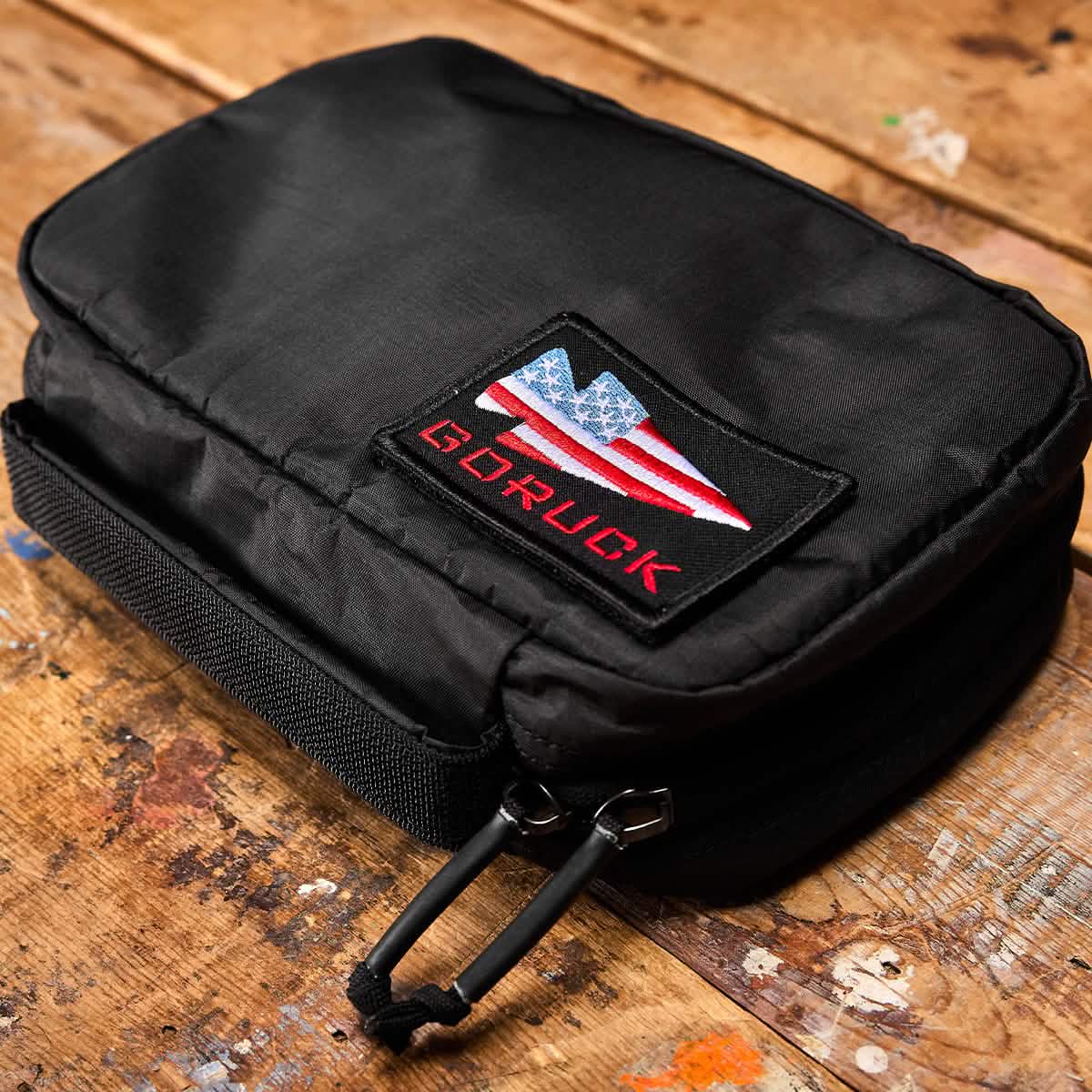 A GR1 Field Pocket - Dyneema, a black rectangular pouch made by GORUCK from durable Dyneema material featuring a double zipper, rests on a wooden surface. It showcases a patch with the American flag and the text "GORUCK" in red, making it an ideal travel accessory for organizing electronics or other essentials.
