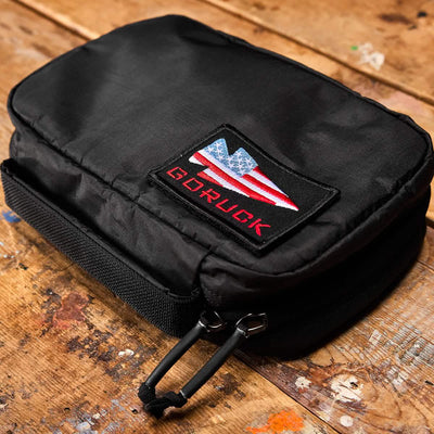 A GR1 Field Pocket - Dyneema, a black rectangular pouch made by GORUCK from durable Dyneema material featuring a double zipper, rests on a wooden surface. It showcases a patch with the American flag and the text "GORUCK" in red, making it an ideal travel accessory for organizing electronics or other essentials.
