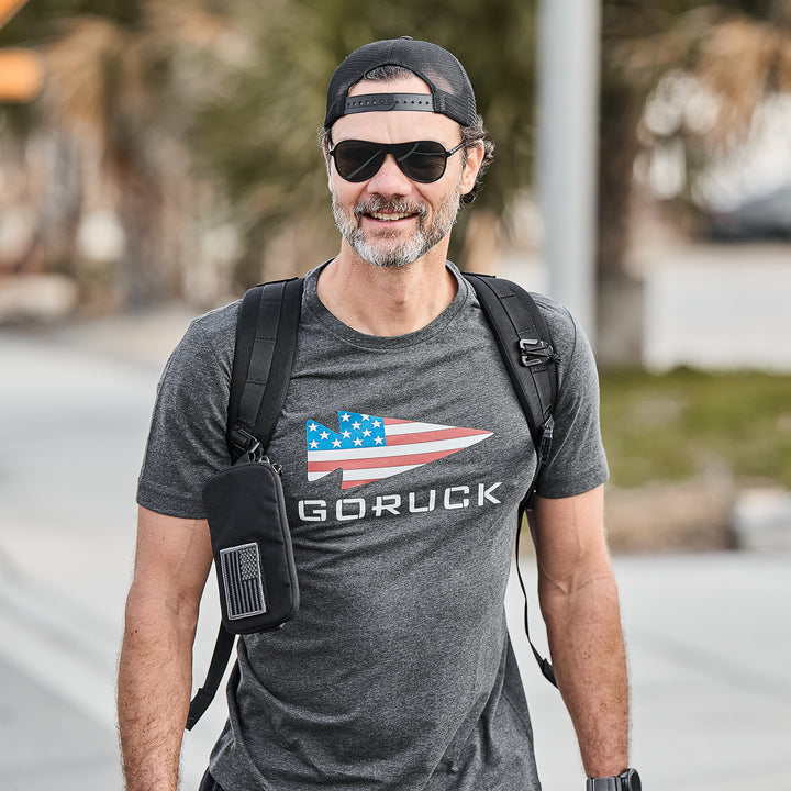 Man wearing sunglasses, a backward cap, and the GORUCK Flag Spearhead Tee - Tri-Blend with a backpack, smiling outdoors.
