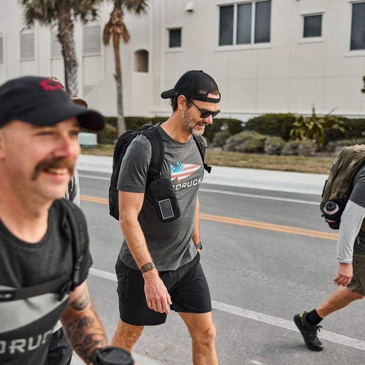 Three men dressed in athletic gear, including GORUCK Flag Spearhead Tee - Tri-Blend shirts and GORUCK backpacks, walk outside near a building and palm trees on a sunny day.