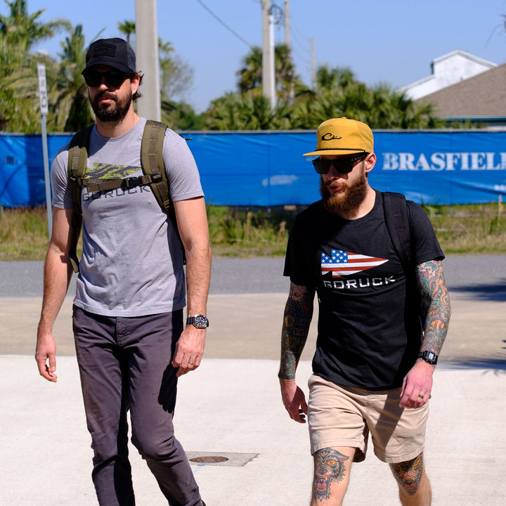 Two men wearing GORUCK USA Tee - Tri-Blend shirts and sunglasses walk outside near a blue construction fence, channeling Special Forces style.