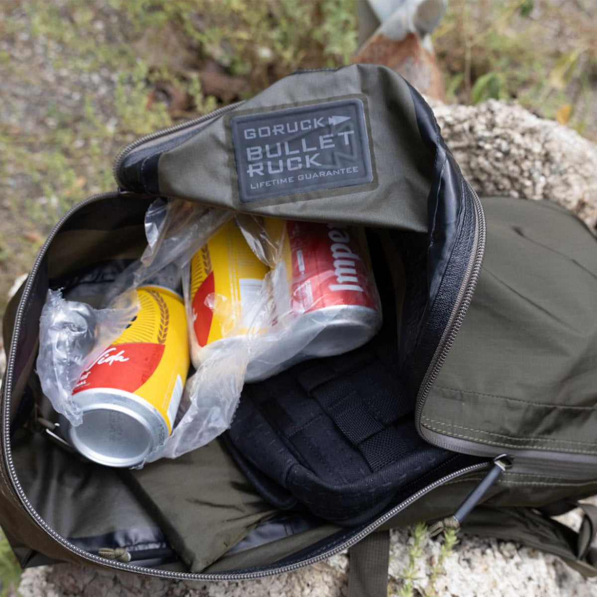 An open GORUCK backpack with cans of soda and a black pouch inside, placed on a rock outdoors.