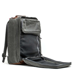 The GR2 - Heritage Waxed Canvas is a gray backpack with brown Red Wing Leather accents, shown open to highlight its interior compartments and mesh pockets—ideal as a stylish travel ruck for any journey.