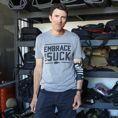 A man in a gray "Embrace the Suck" shirt stands before shelves of gear, smiling and ready to boost mobility with the Recovery Floss Band.