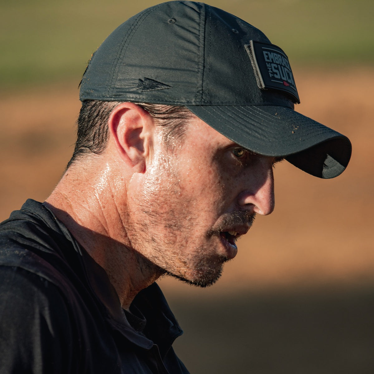 A man sweating heavily wears a black cap and shirt outdoors in bright sunlight, his Performance TAC Hat - TOUGHDRY designed to handle rugged conditions.