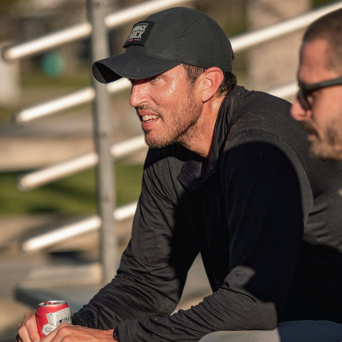 A sweaty man in a black shirt and a Performance TAC Hat - TOUGHDRY sits outdoors with a drink beside another person.