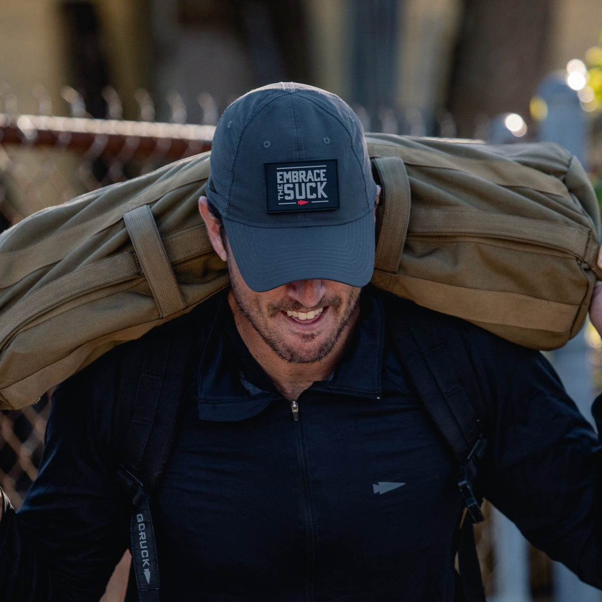 A man wearing the Performance TAC Hat - TOUGHDRY carries a large tan duffel bag on his shoulders outdoors, with a fence in the background.