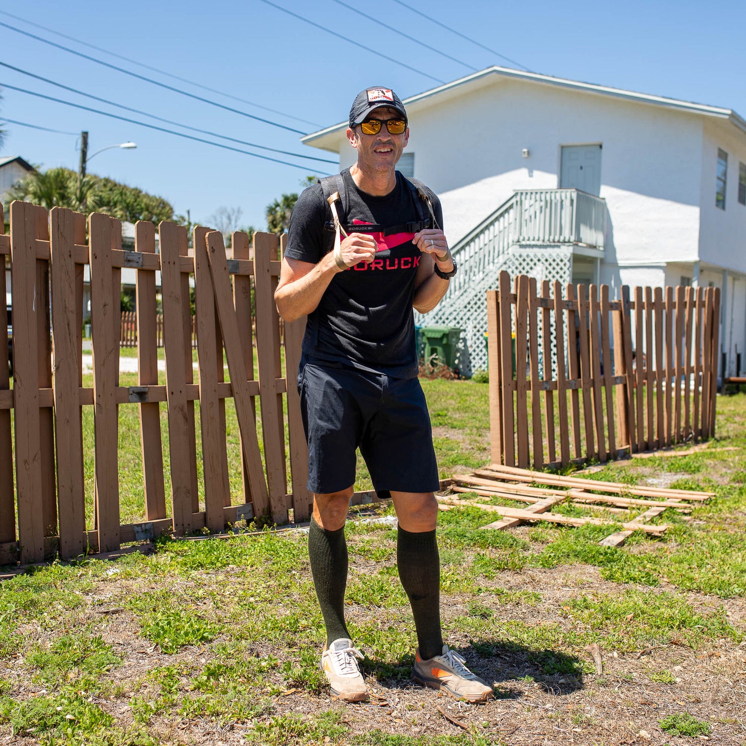 Wearing athletic gear, a man with T-REX Hand Straps and a drink stands by a broken wooden fence on a sunny day, protecting his elbow from tendinitis.