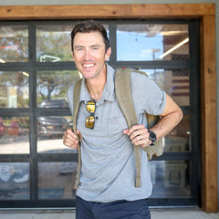 A smiling man wearing sunglasses and a gray polo stands in front of a garage door with the GR2 - Heritage Waxed Canvas backpack, featuring a durable laptop compartment.