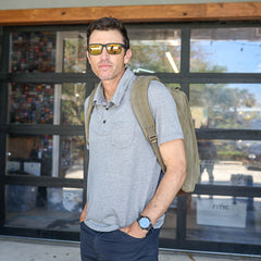 A man wearing sunglasses and a gray polo stands outside a building with glass doors, carrying the GR2 - Heritage Waxed Canvas ruck, ready for adventure.
