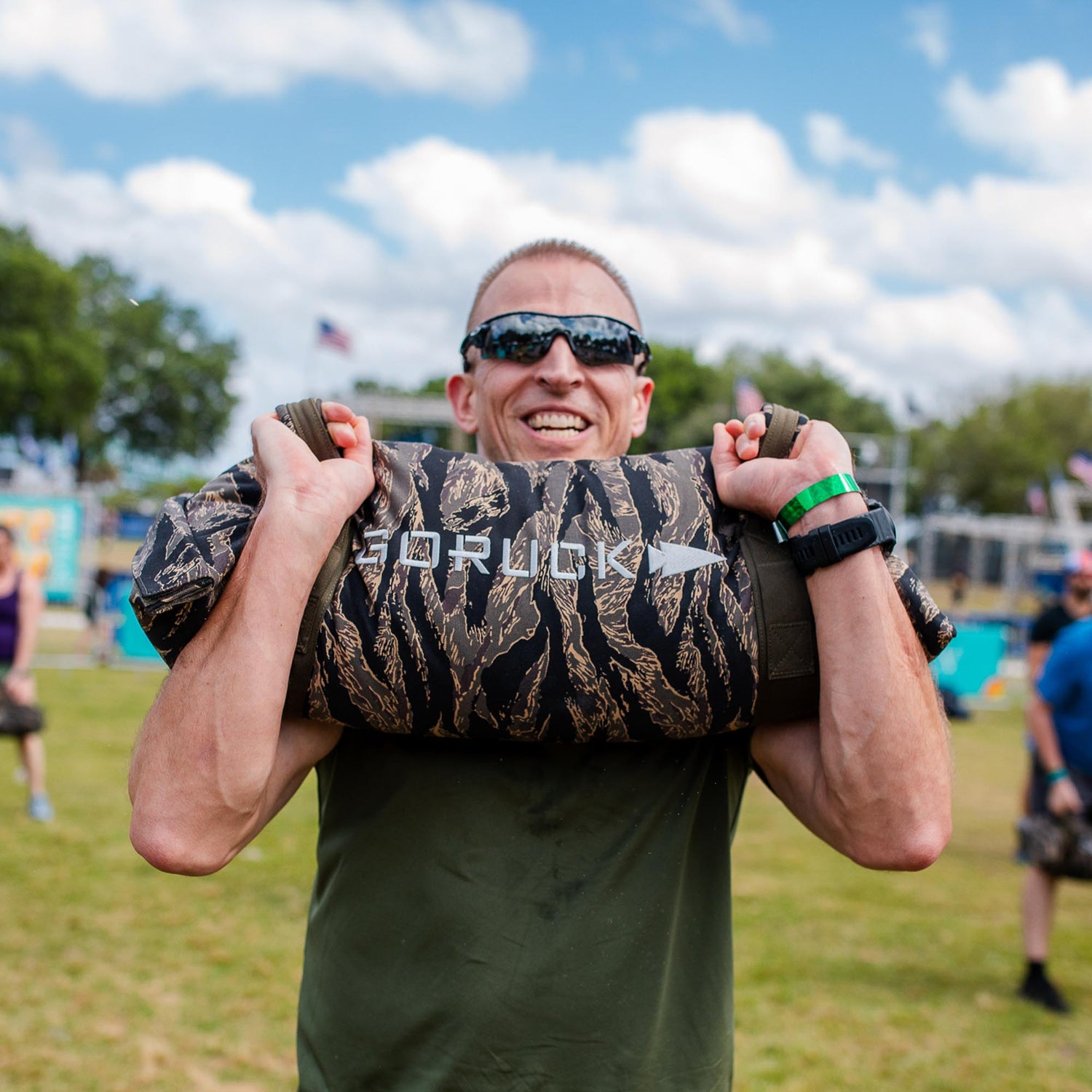 A smiling man in sunglasses and a green shirt lifts Simple Training Sandbags outdoors, demonstrating effective sandbag workouts beyond the home gym.