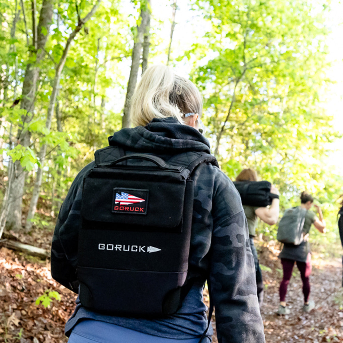A group hikes a forest trail with black RPC 3.0 + 10lb Ruck Sandplate carriers for weighted training beneath bright green trees.