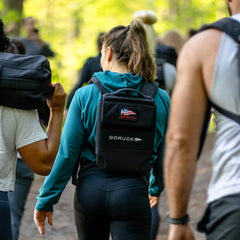 A group hikes outdoors, one carrying the black RPC 3.0 + 10lb Ruck Sandplate backpack with an American flag patch—ideal for weighted training and rucking challenges.