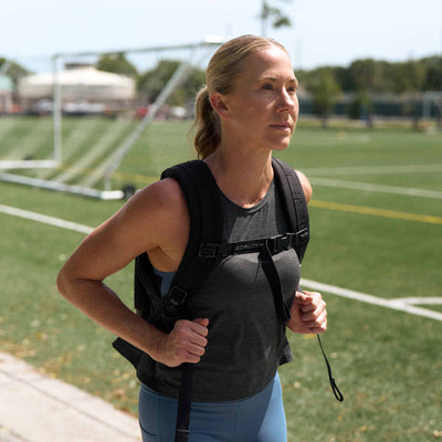 A woman with a backpack walks on a sunny sports field, wearing the Women’s USA Performance Tank – ToughMesh for ultimate comfort.