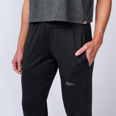 A person wearing black Women’s Performance Joggers made from performance fabric and a gray top, with one hand in a pocket, is shown from waist to knee.