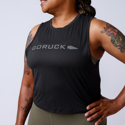 A smiling, tattooed woman wears a black Women’s USA Performance Tank - ToughMesh, featuring featherweight ToughMesh fabric, and olive leggings with a hand on her hip.