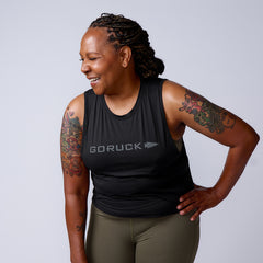 A smiling woman with braided hair and tattoos wears the Women’s USA Performance Tank – ToughMesh by GORUCK in black, paired with olive leggings.