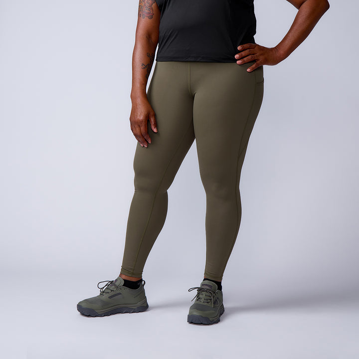 A woman wearing the Women’s Training Leggings Pocket - ToughFlex in olive green, paired with a black top and green shoes, stands with one hand on her hip against a plain background.