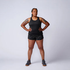 Fit woman with tattoos wearing black athletic tank top, camo shorts, and gray sneakers standing confidently