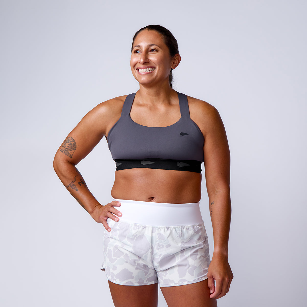 A woman smiles in ToughStretch Women’s Training Shorts and a sports bra, standing with one hand on her hip against a plain background.