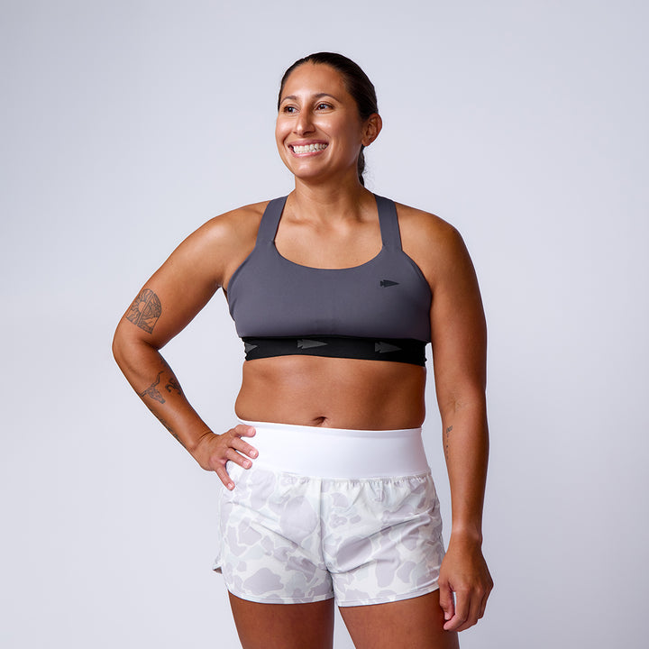 A woman smiles in ToughStretch Women’s Training Shorts and a sports bra, standing with one hand on her hip against a plain background.