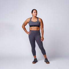 Fit woman wearing gray athletic leggings, matching sports bra, and gray trail shoes, standing against plain background