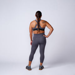Back view of woman in gray sports bra, leggings, and sneakers standing against white background, sporting tattoos
