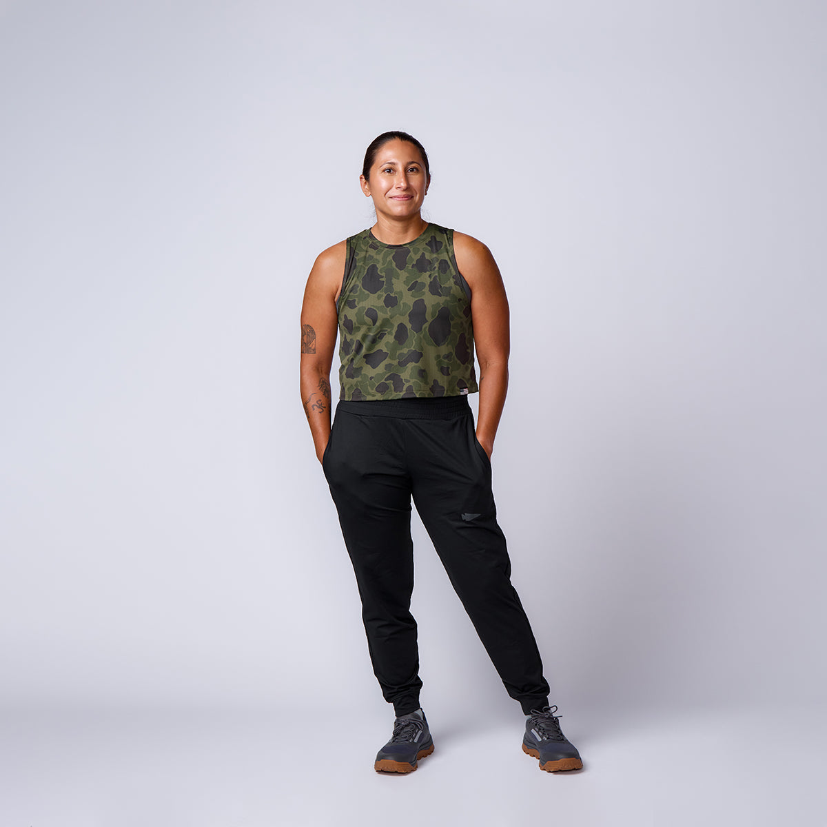 A woman stands smiling, wearing the Women’s USA Performance Tank - ToughMesh in green camo, paired with black joggers and grey sneakers.
