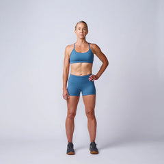 Fit woman in blue sports bra and shorts standing confidently on plain studio background wearing black sneakers