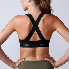 A woman is shown from behind wearing the Power Bra - Toughflex, a black cross-back, high-impact sports bra, paired with olive green leggings.