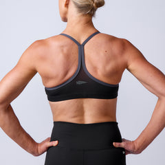 A woman is shown from behind wearing the Stealth Bra - ToughFlex, a minimalist sports bra with lightweight support, paired with high-waisted leggings and her hands on her hips.