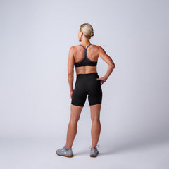 A woman in athletic wear stands with her back to the camera, hand on hip, highlighting the Women’s Biker Shorts - ToughFlex against a plain light background.
