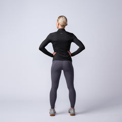 Woman standing with hands on hips wearing black long-sleeve athletic top and gray leggings on plain background