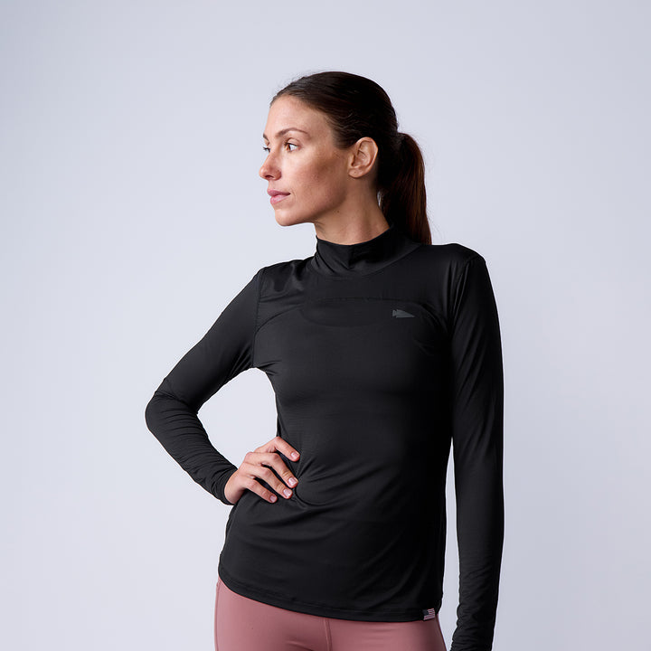 Woman in a black Women’s Long Sleeve Mock Neck - ToughMesh athletic top with UPF protection and pink leggings stands with hand on hip, looking to the side.