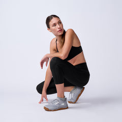 Woman in black sports bra and leggings crouching wearing gray GORUCK rucking shoes on white background