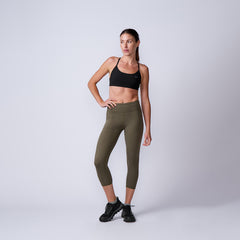 Fit woman in black sports bra and olive green cropped leggings posing against plain gray background