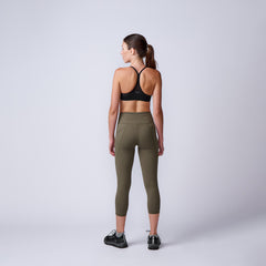 Woman in black sports bra and olive green leggings with sneakers standing against plain backdrop