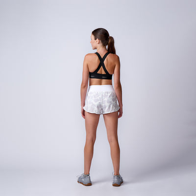 A woman stands with her back to the camera, wearing a black sports bra, Women’s Training Shorts - ToughStretch, and gray sneakers against a plain background.