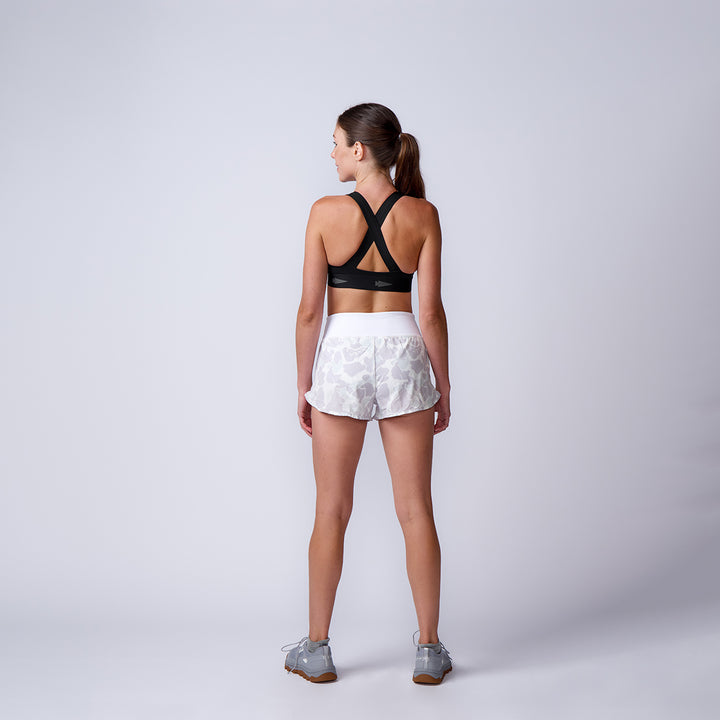 A woman stands with her back to the camera, wearing a black sports bra, Women’s Training Shorts - ToughStretch, and gray sneakers against a plain background.