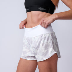 A woman adjusts the waistband of Women’s Training Shorts - ToughStretch, featuring a black sports bra and white patterned shorts for a secure, comfortable fit.